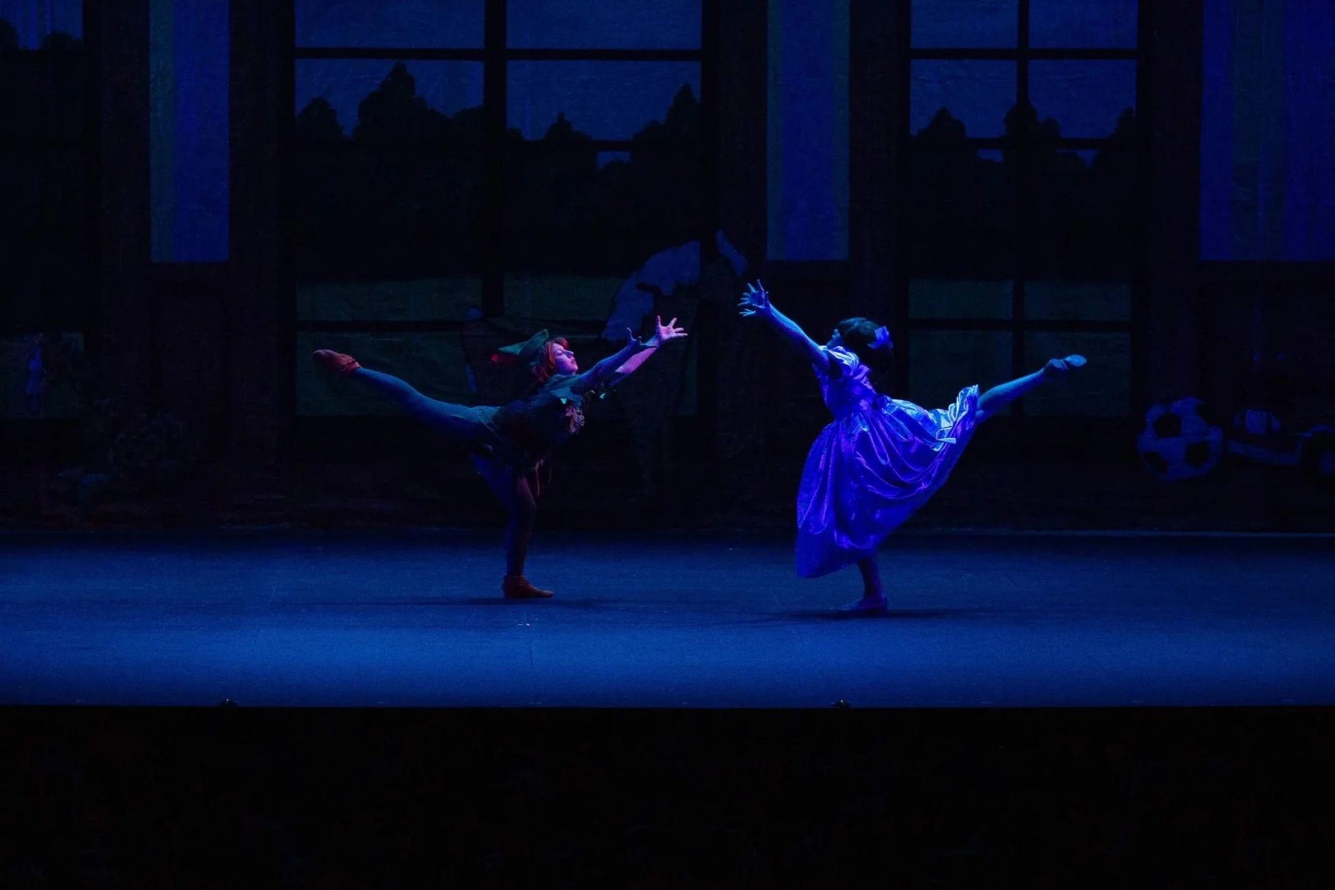 Two ballet dancers perform on stage in dim lighting, one dancer is wearing a black dress and the other a purple dress, both executing a high leg kick with their arms extended.