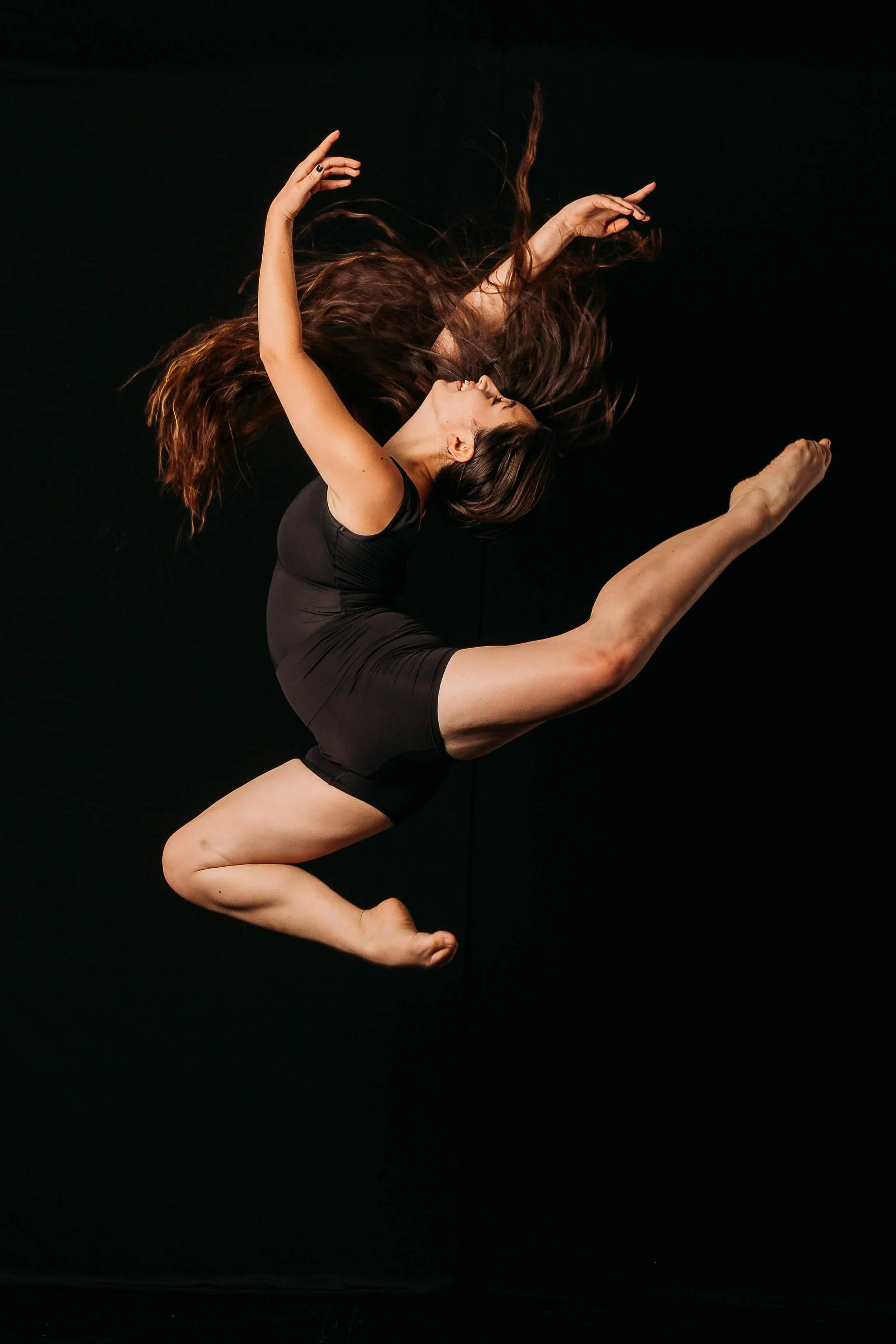 A woman with long brown hair performing a dance pose on a black background, wearing a black athletic outfit.