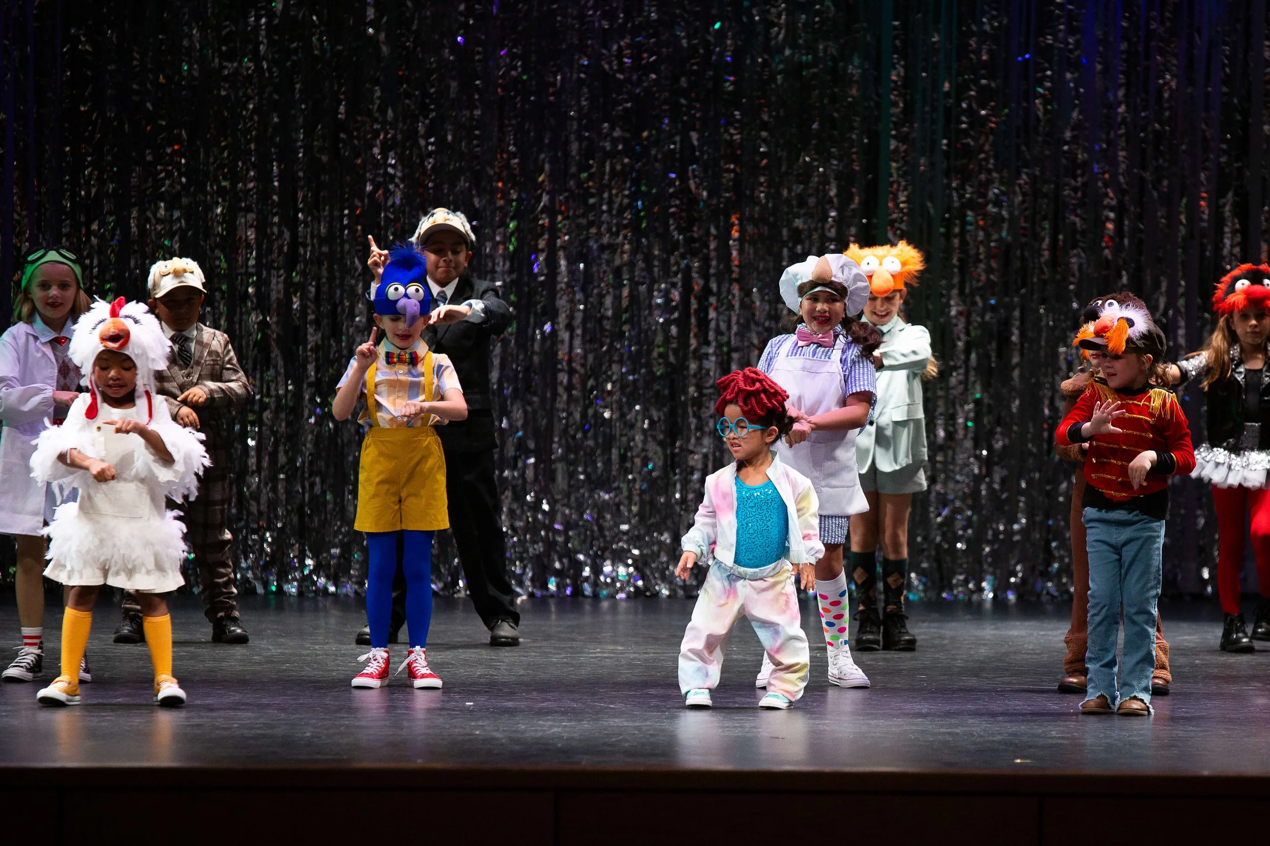 Children dressed in colorful costumes performing a dance on stage with a glittering black backdrop.