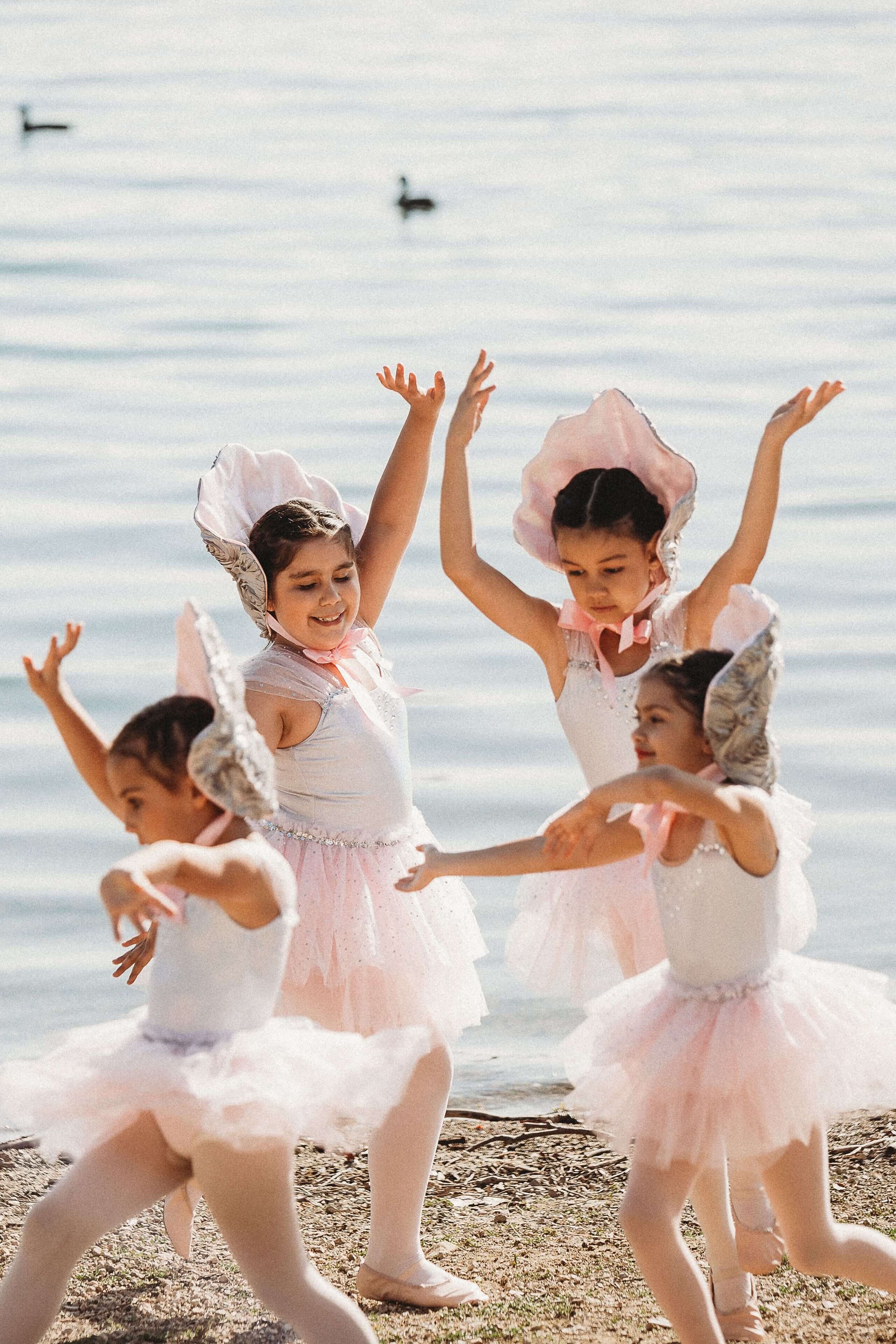 Children in ballet costumes dancing on a beach with water in the background.