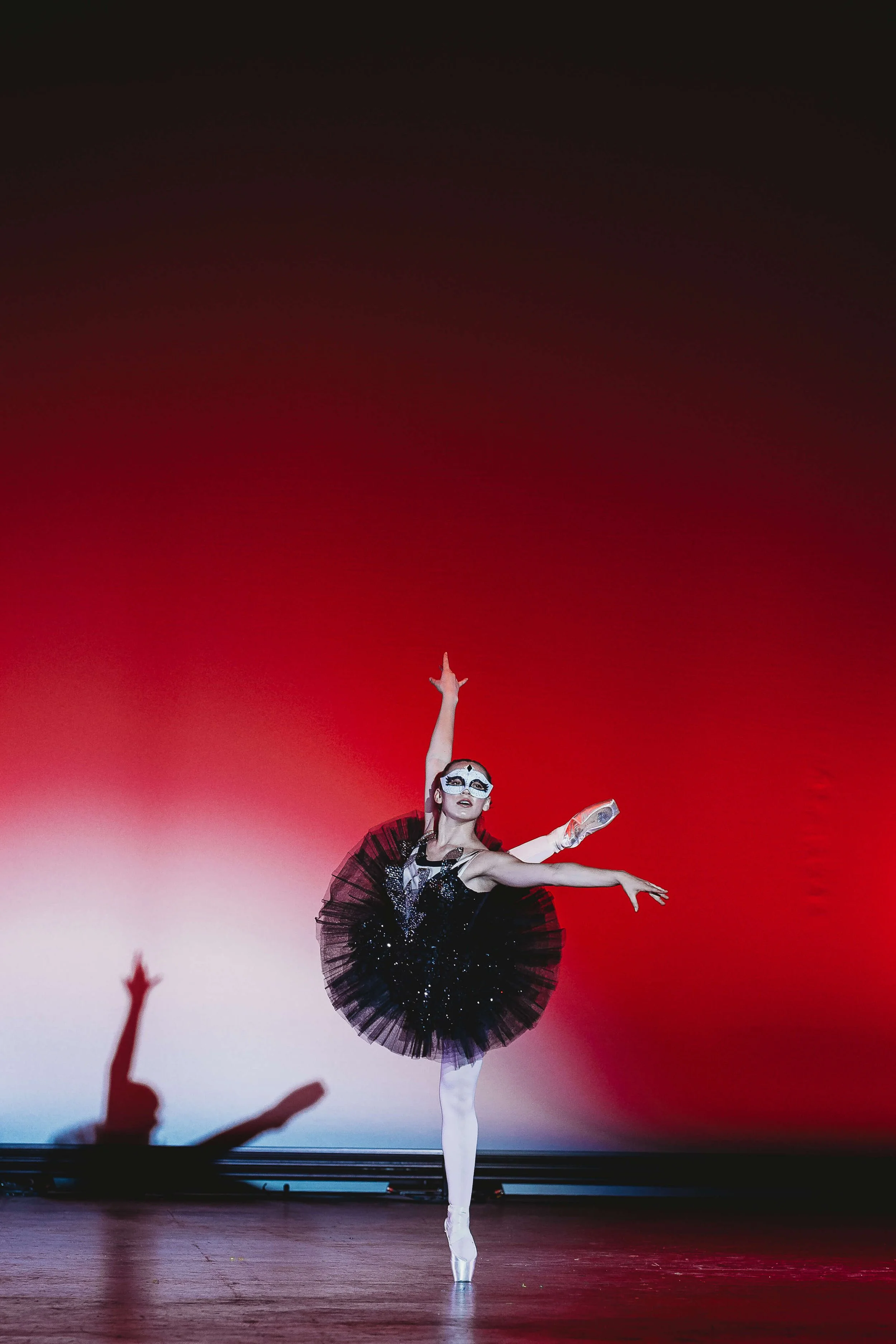 Ballet dancer performing on stage in a black tutu and white tights, with makeup resembling a skull or skeleton, against a red gradient background, with a shadow of a hand on the floor nearby.