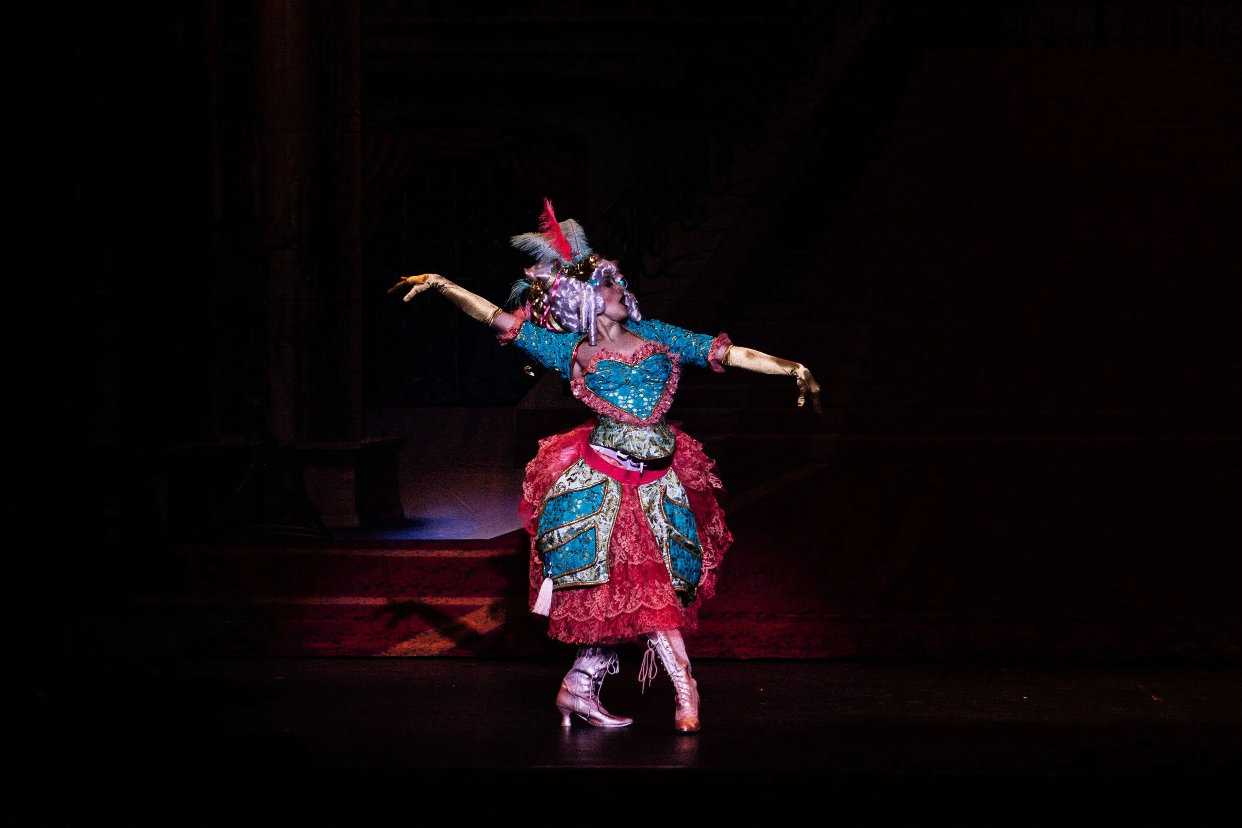 Performer in colorful elaborate costume, in a dance pose on stage with dark background and spotlight.