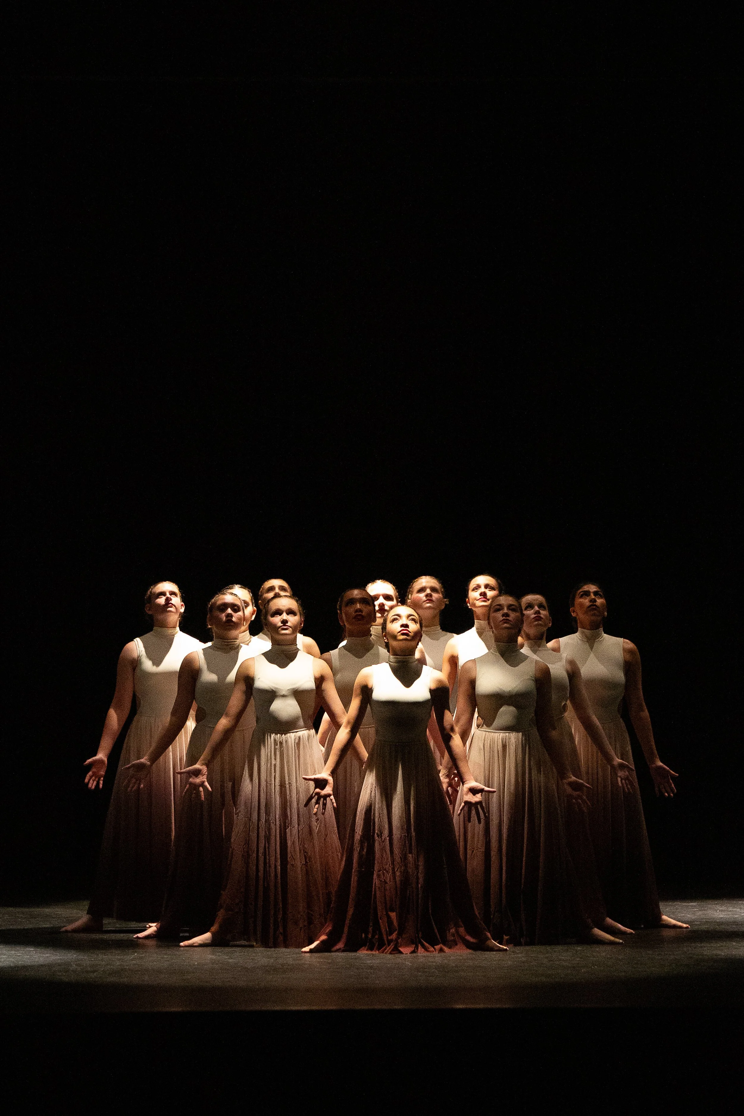 A group of female ballet dancers in white costumes standing on stage, illuminated from below, against a black background.