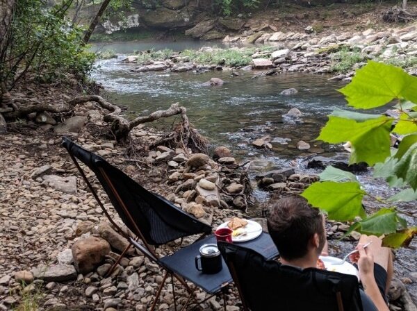 man sitting in camp chair by river eating easy camping meal drinking coffee