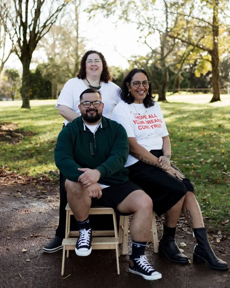 Three people sitting and standing outdoors on a grassy area with trees in the background, smiling at the camera. One person is sitting on a small wooden stool, wearing glasses, a green jacket, shorts, and sneakers. The woman next to him is sitting on a larger stool, wearing a white T-shirt with red text, black skirt, and black boots. The third person is standing behind them, wearing glasses, a white top, and smiling.