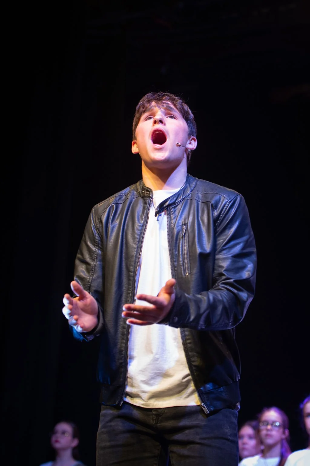A young man speaking or singing passionately on stage, wearing a black leather jacket and white shirt, with a dark background and some people in the background.