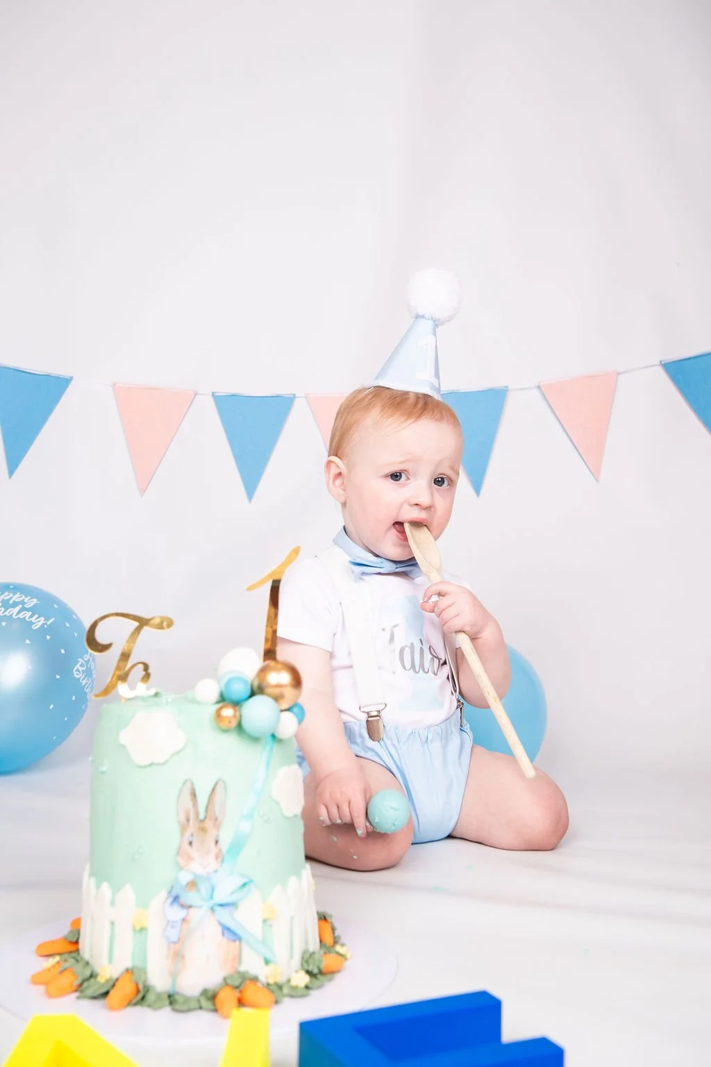 A young boy at a birthday party, sitting on the floor, wearing a party hat and holding a wooden spoon. There is a decorated birthday cake with an Easter bunny theme, balloons, and colorful triangular banners in the background.