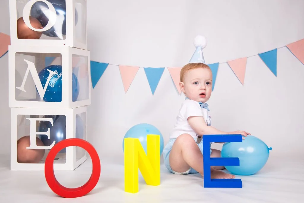 A baby wearing a party hat sitting on the floor with balloons and colorful letters spelling 'ONE' for a first birthday celebration, with a backdrop of pennant banners and a gift box with more balloons inside.
