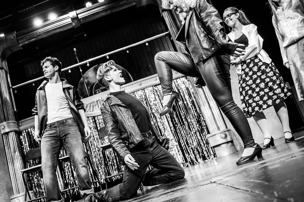 Group of people performing on a stage, some standing and one kneeling, with a woman in leather pants being lifted.