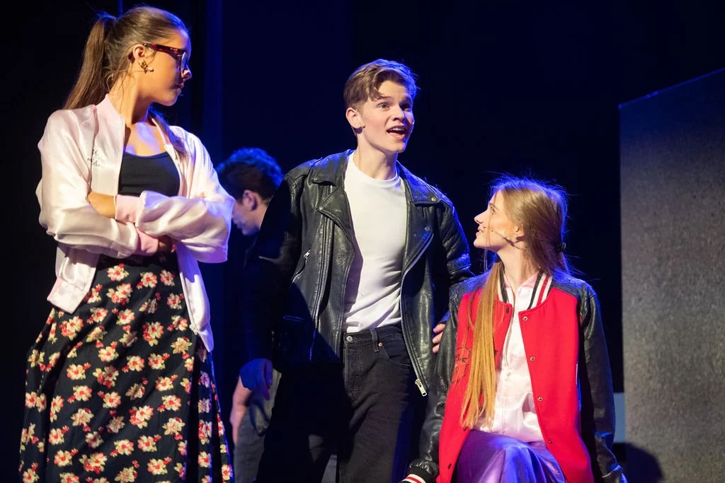 Three teenagers on stage during a performance: a girl with glasses and a floral skirt on the left, a smiling boy in a black leather jacket and white T-shirt in the center, and a girl with long hair in a red and black jacket sitting on a chair on the 