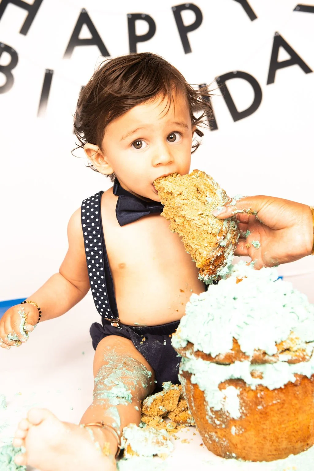 A young boy with brown hair, wearing a black bowtie and polka dot suspenders, is being fed a large piece of cake during a birthday celebration. There is a 'Happy Birthday' banner in the background, and the boy is surrounded by cake and blue frosting.