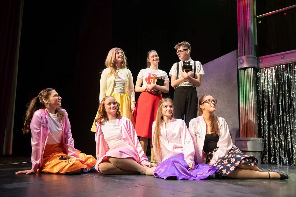 Group of young performers dressed in colorful 1950s style costumes on stage, some sitting and others standing, during a theatrical performance.