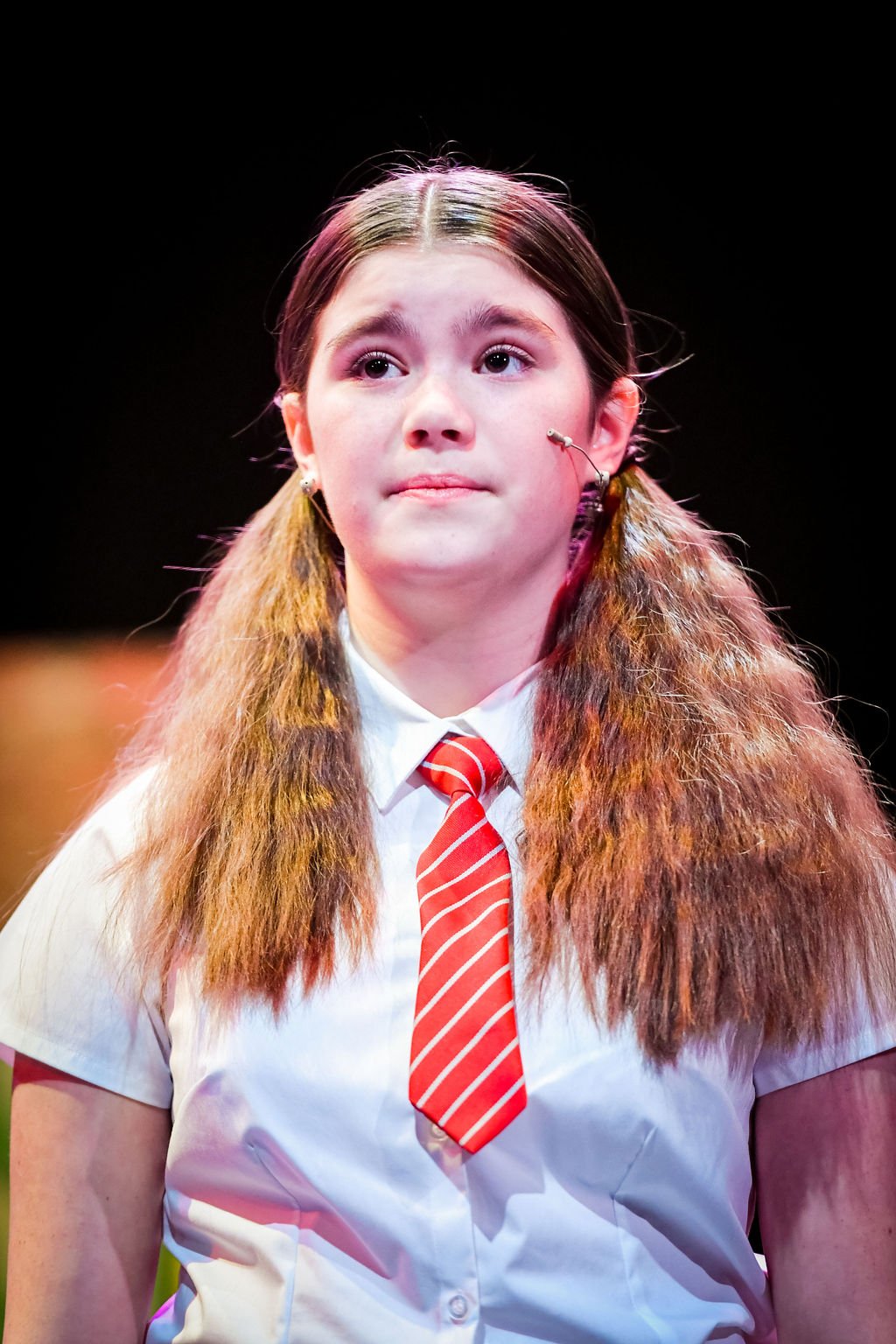 Young girl with long, wavy brown hair, wearing a white shirt, red striped tie, and using a microphone headset, on a stage with a dark background.