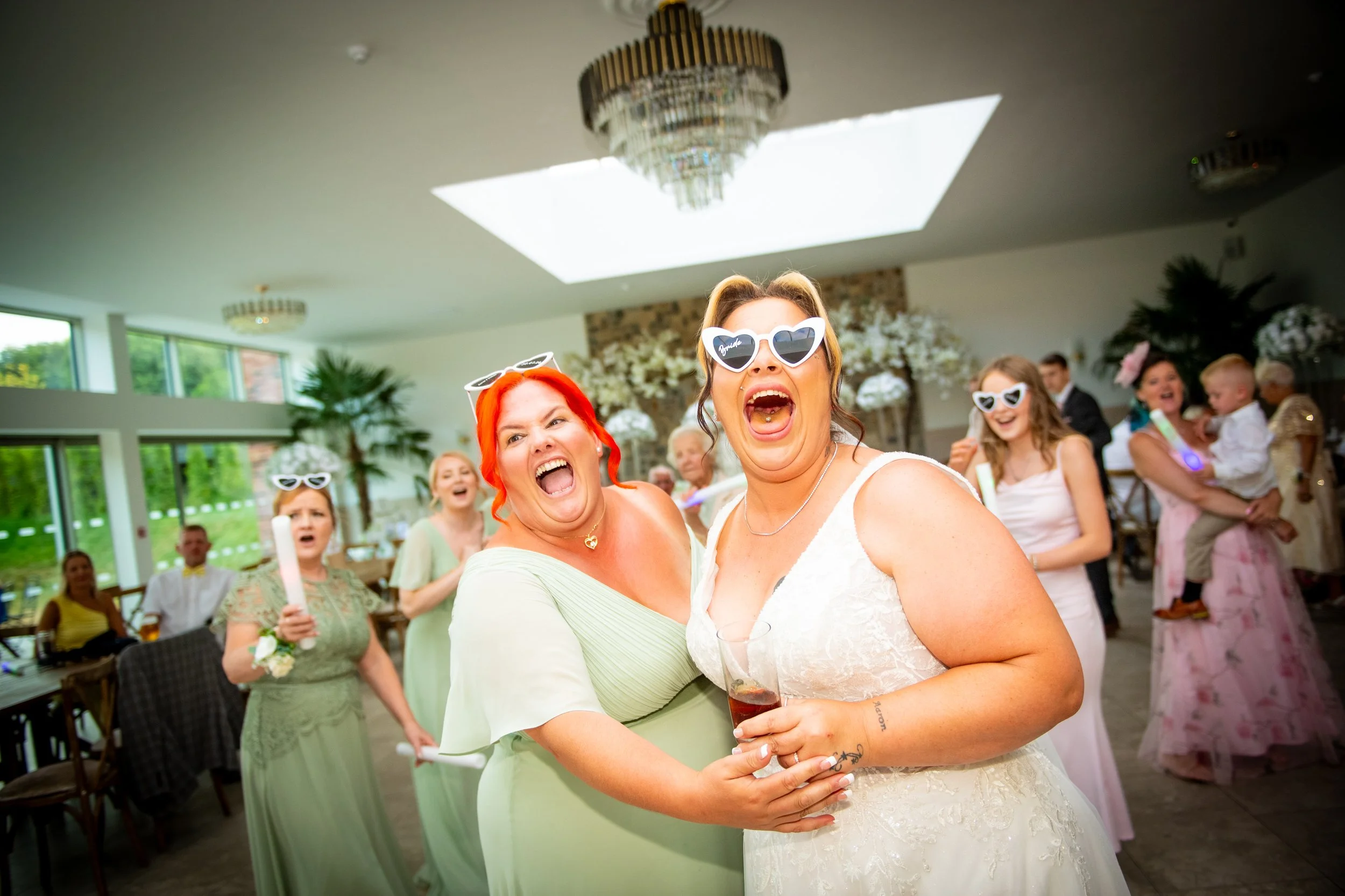 Bride and bridesmaids wearing white heart sunglasses laughing during high-energy wedding reception