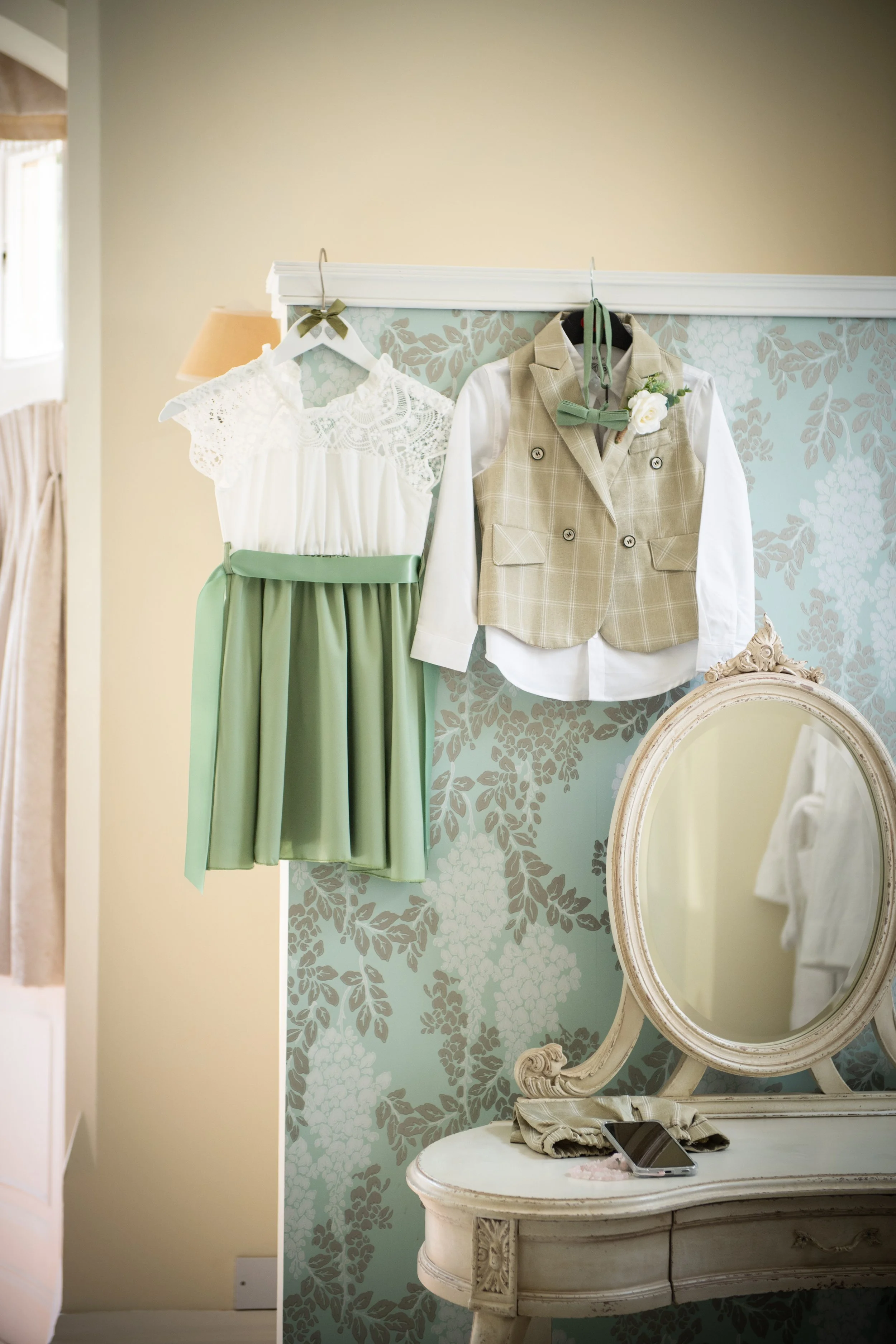 Bridal party outfits in sage green and beige hanging neatly above a vintage vanity table