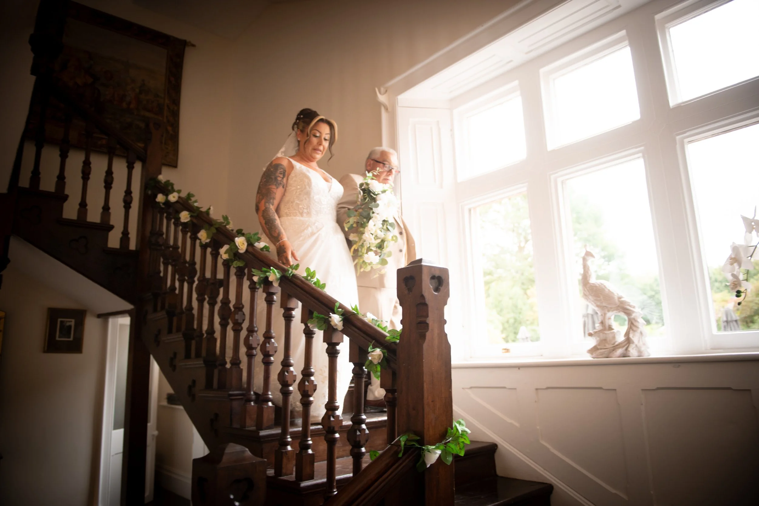 Bride descending the grand wood staircase at Cefn Tilla Court bathed in natural window light