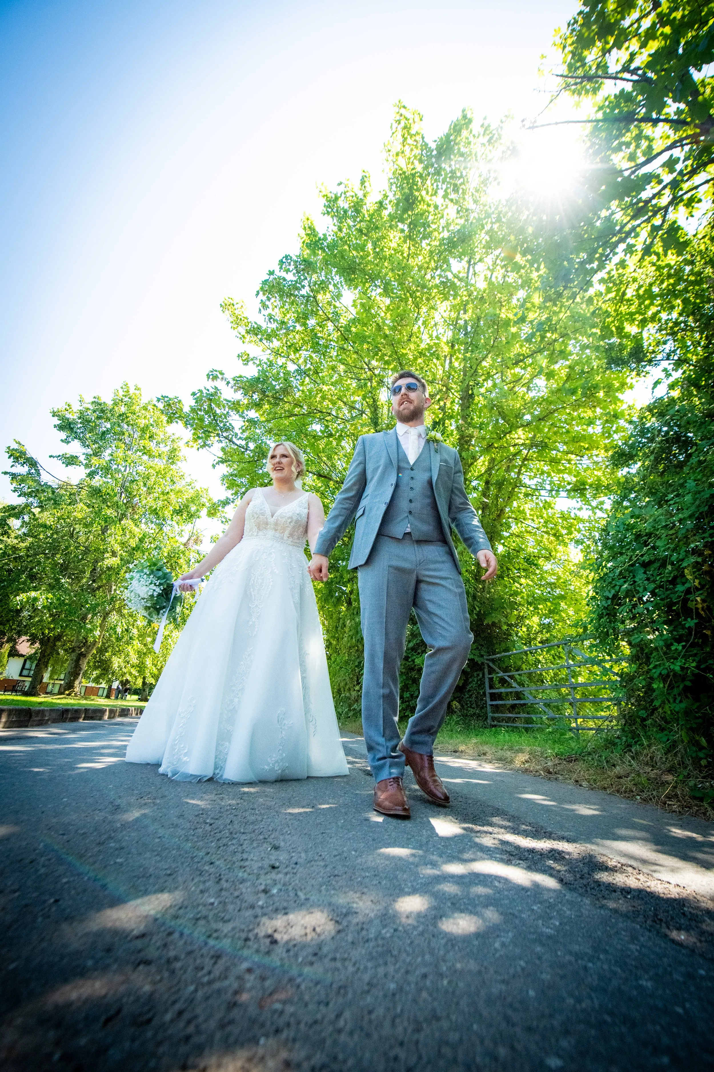 Bride and groom wearing sunglasses walking together under green trees during a hot summer wedding in South Wales