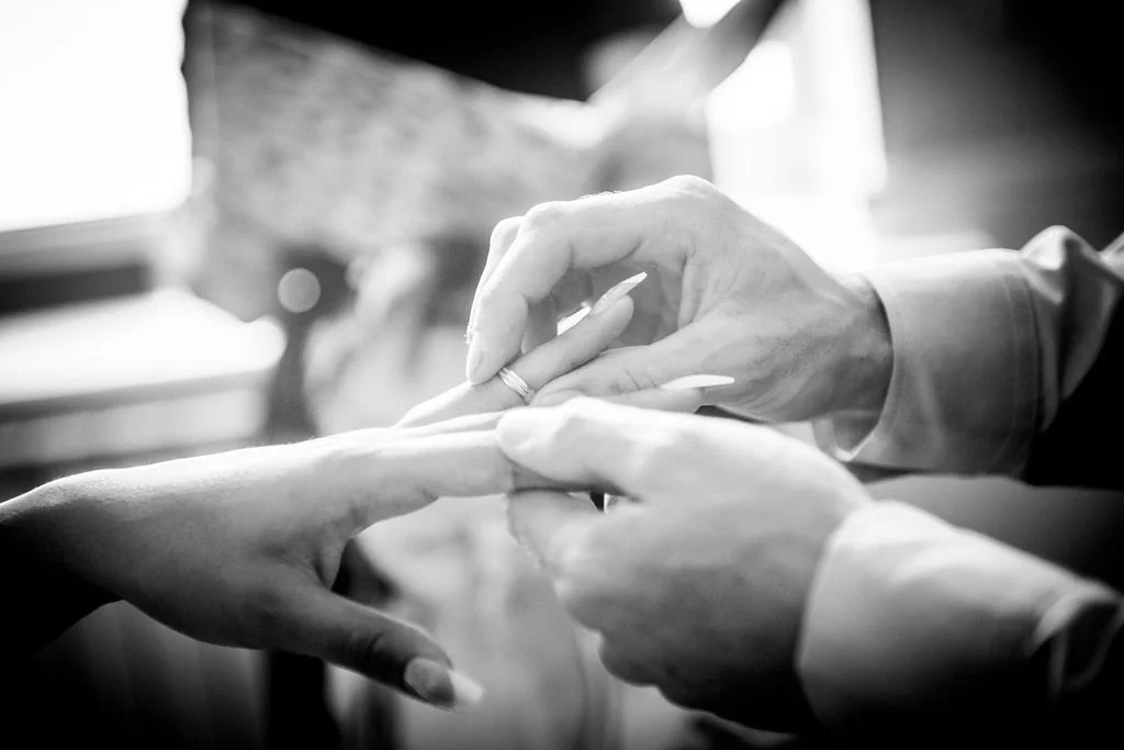 Black and white close up of a groom placing a wedding ring on his bride's finger