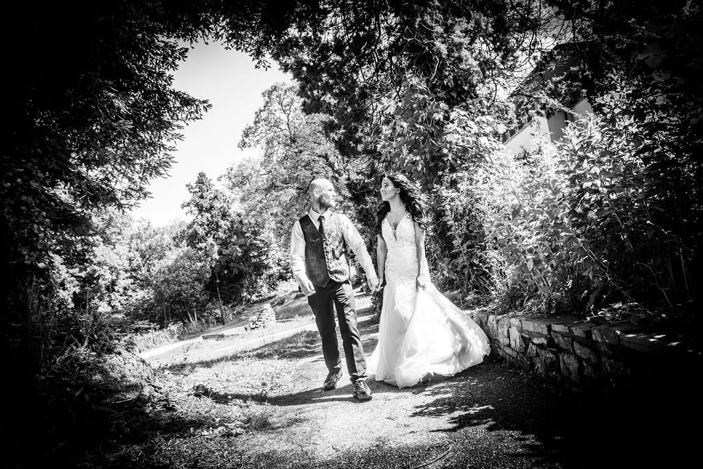  Black and white photo of a bride and groom walking together under trees in South Wales
