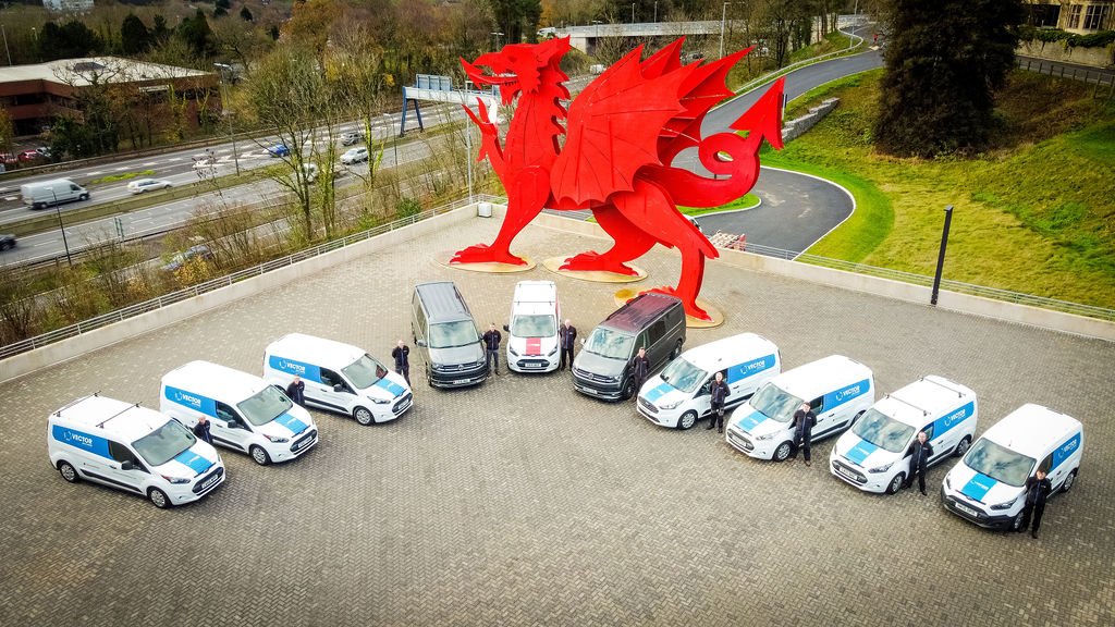 A large red dragon sculpture is situated behind a fleet of white and gray service vans parked in a curved formation on an open paved area.