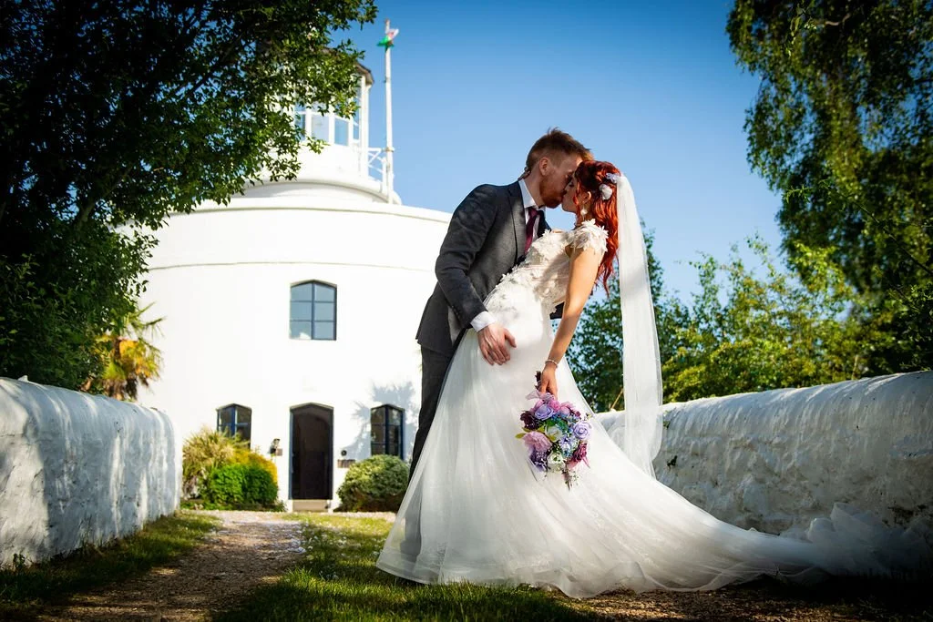 A romantic portrait of a groom dipping his bride for a kiss on the pathway outside West Usk Lighthouse, photographed by Emotion Picture Photography.
