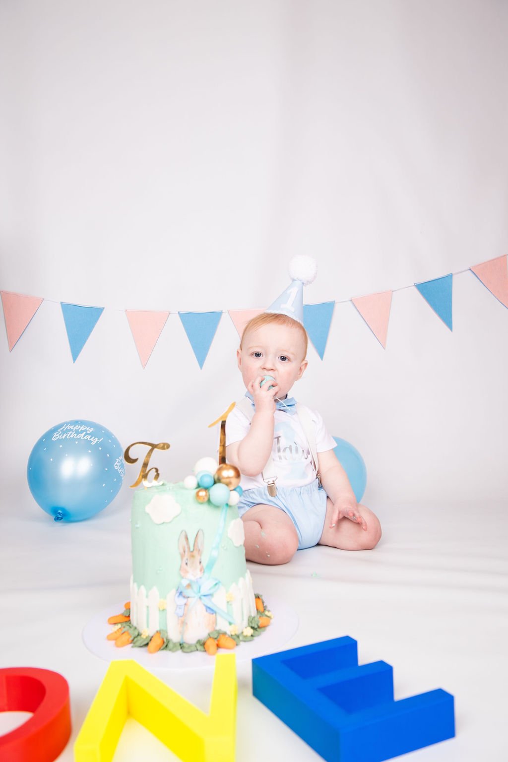 A young boy celebrating his first birthday with a cake, balloons, and colorful decorations. He wears a party hat and grips a cake with a bunny and clouds design. There are letters and balloons in the background.