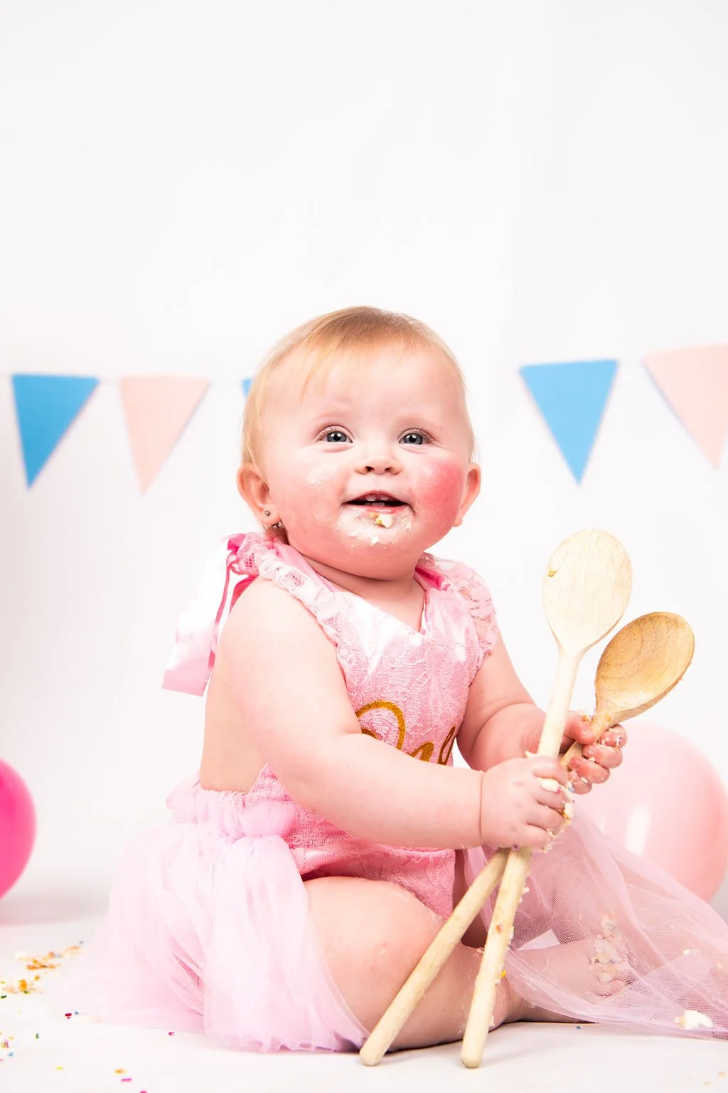 A smiling baby girl in a pink dress sitting on the floor with cake crumbs on her face, holding two wooden spoons, with pink and blue bunting flags in the background, celebrating her birthday.