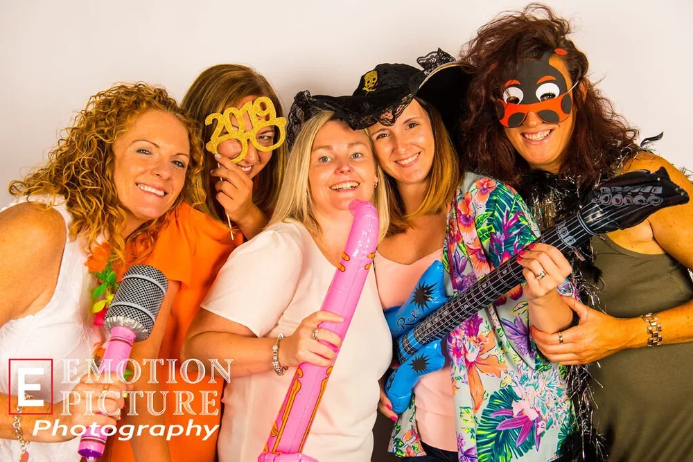 Group of five women celebrating New Year's Eve, wearing colorful costumes, sunglasses, and holding inflatable party props, with a plain background.
