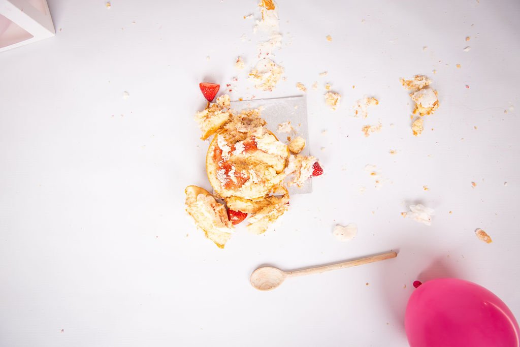 A broken glass with pieces of cake and strawberries spilled on a white surface, a wooden spoon and a pink plastic cup nearby.