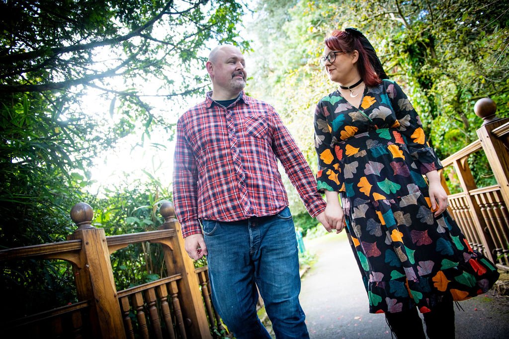 A man and woman holding hands and walking outdoors on a path surrounded by greenery and trees.