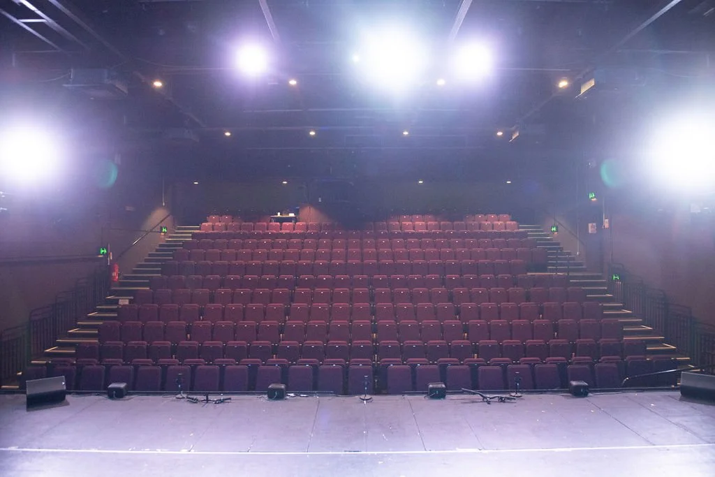 Empty theater with red seats, stage lights overhead, and microphones on the stage floor.