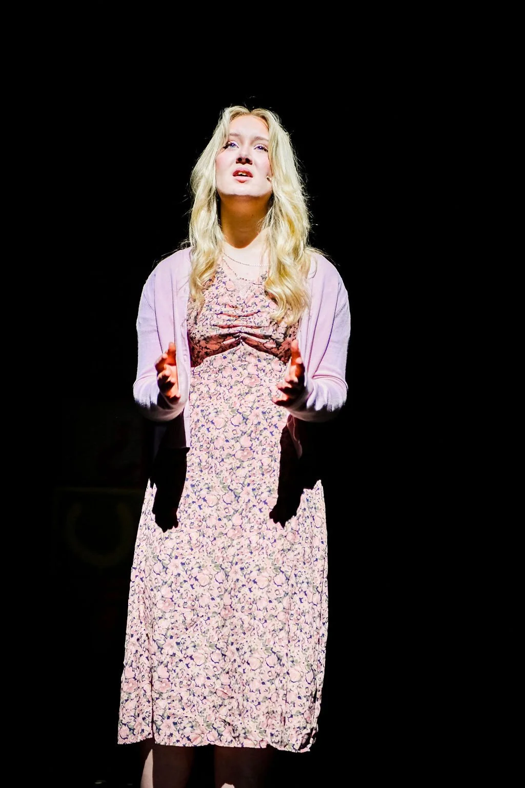 A young woman with blonde hair stands on stage, wearing a floral dress and a light pink cardigan, singing or speaking passionately against a dark background.