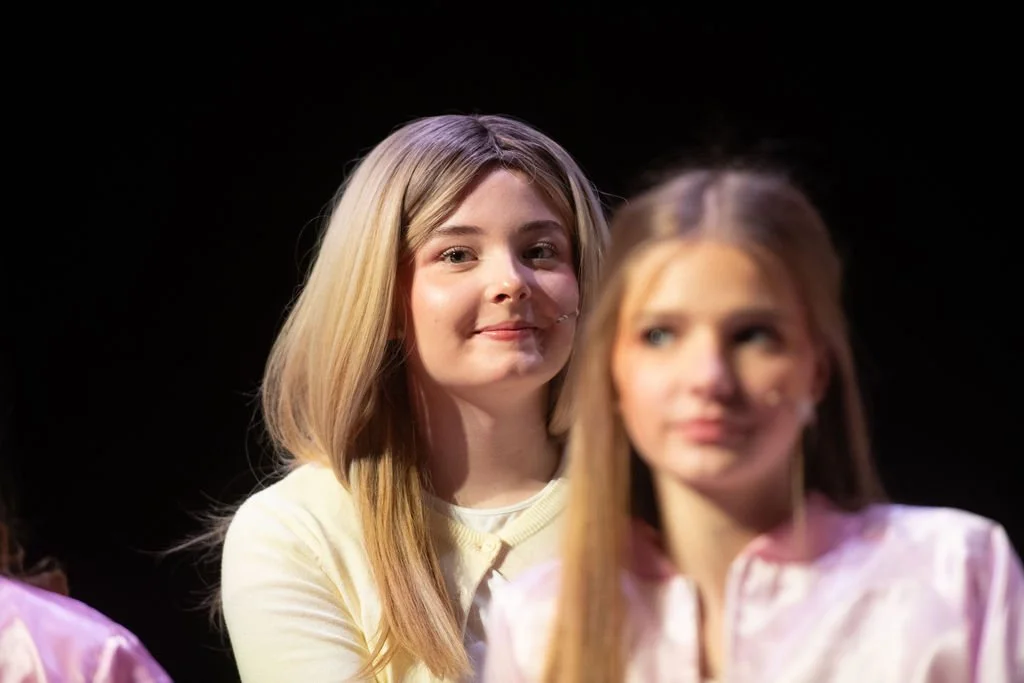 Two young women sitting side by side on stage, with one smiling and the other looking away, against a dark background.