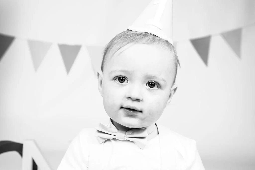 Black and white photo of a young child wearing a party hat and bow tie, with a banner in the background.