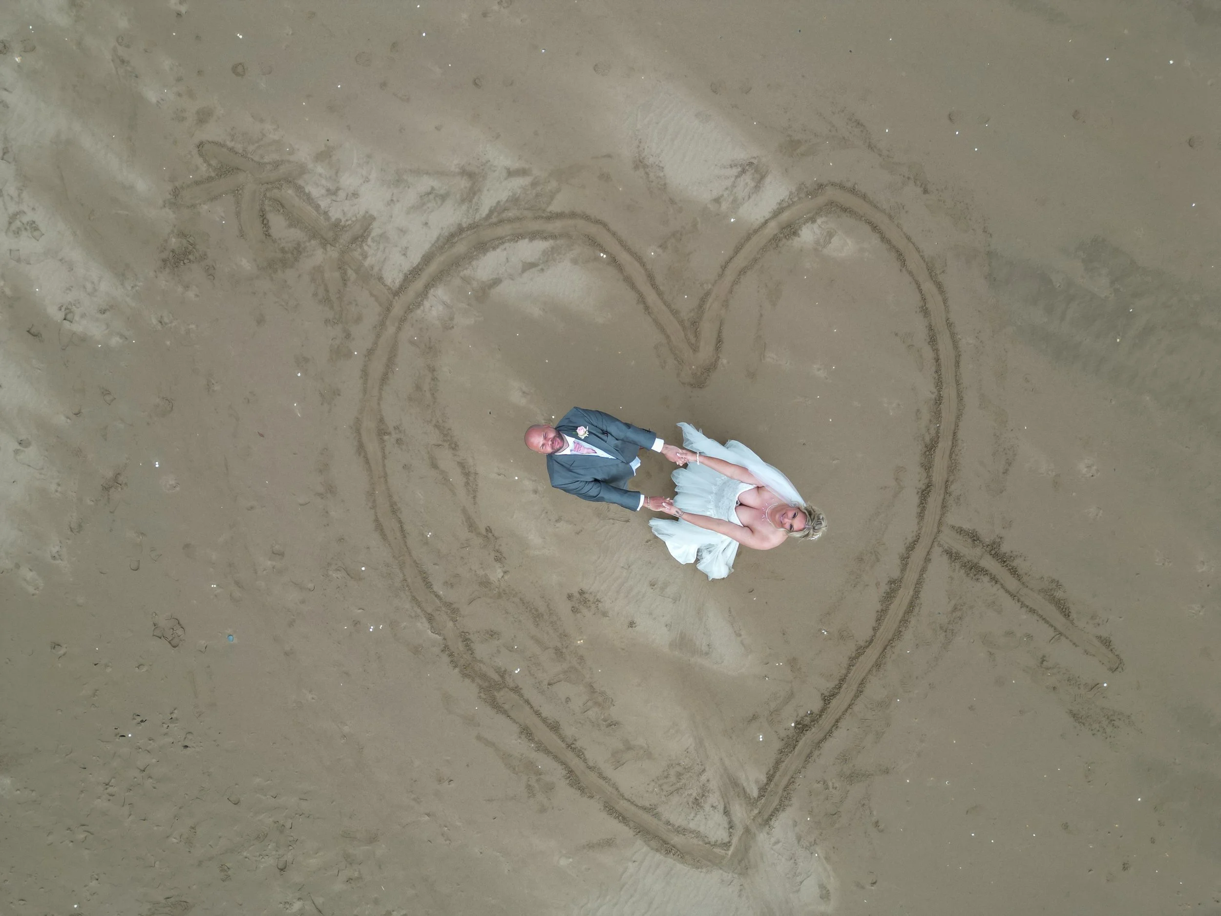 A bride and groom in wedding attire holding hands inside a heart shape drawn in the sand on the beach.