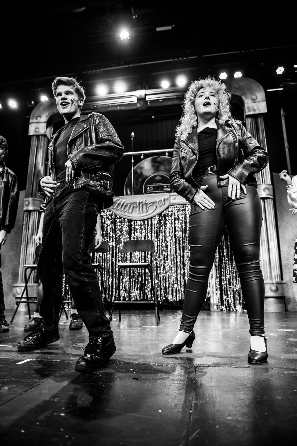 Black and white photo of two performers on stage dressed in leather jackets, with a decorated curtain behind them, and chairs and a drum set in the background.