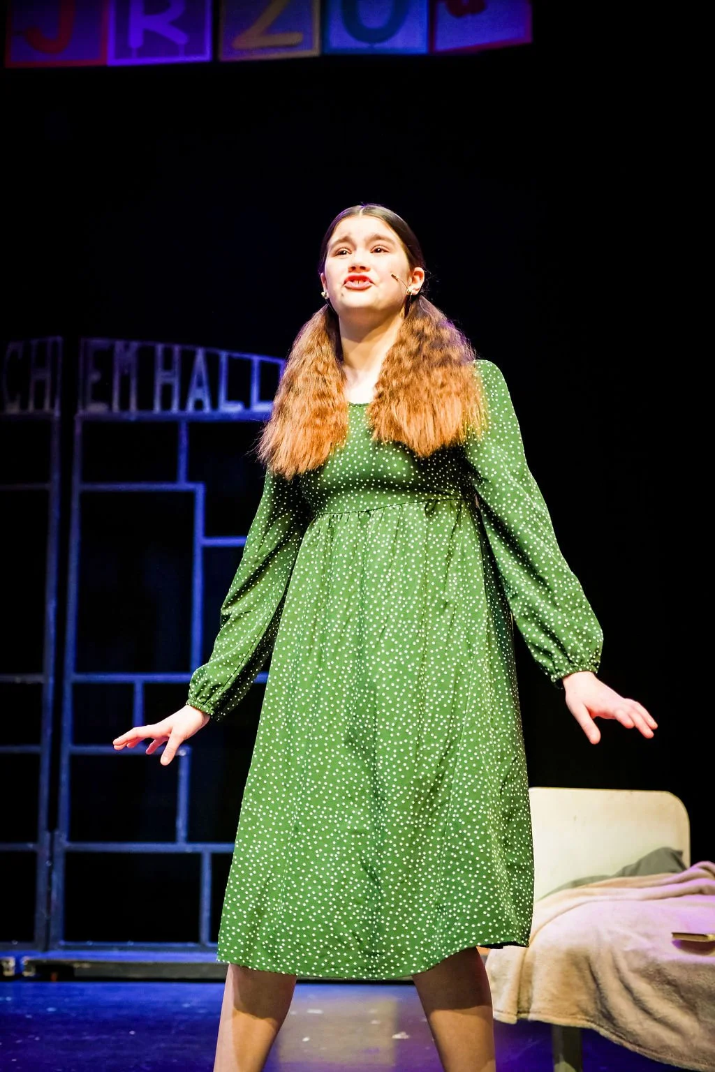 Young woman performing on stage wearing a green dress with white polka dots, standing in front of a black background with stage props.