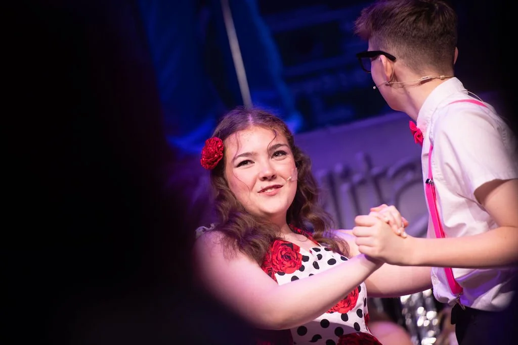 A girl with long curly hair and a red flower hair accessory, wearing a dress with a red and black rose pattern and black polka dots, is smiling as she dances while holding hands with a boy in glasses, a white shirt, suspenders, and a pink bow tie.