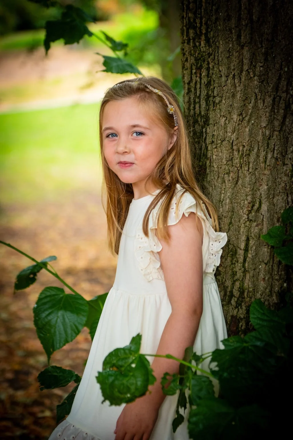 Color portrait of a young girl in a white dress leaning against a large tree during a family photoshoot at Belle Vue Park in Newport by Emotion Picture Photography.