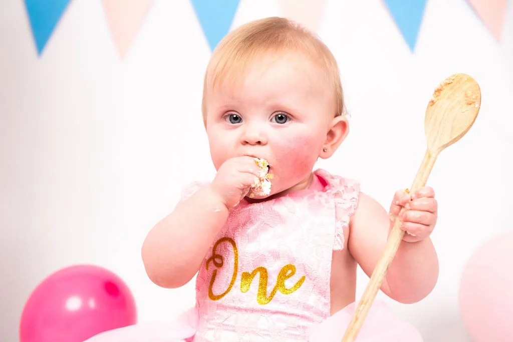 A young girl celebrating her first birthday, wearing a pink dress with a "One" bib, holding a wooden spoon, and surrounded by pink and blue balloons with pastel-colored bunting in the background.