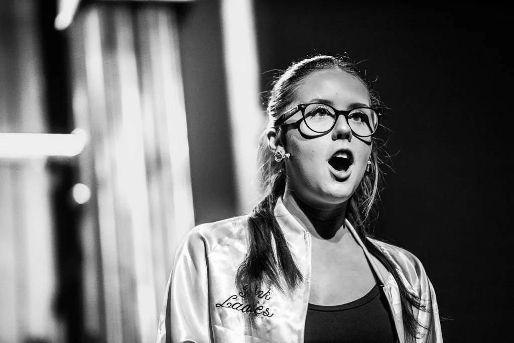 A young woman with glasses, dark hair in pigtails, wearing a jacket with embroidered text, appears to be singing or speaking on stage with an expressive face.