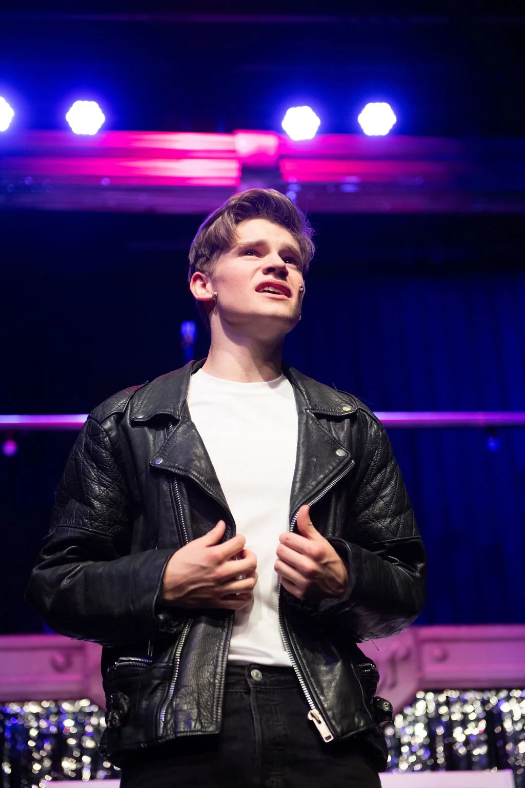 A young man with a pierced eyebrow wearing a leather jacket and white t-shirt stands on stage with purple and pink lighting behind him, showing an emotional expression.