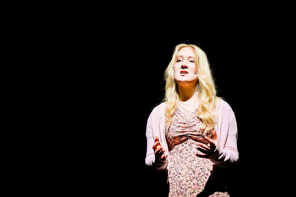 Young woman with blonde curly hair performing on stage in front of a dark background.