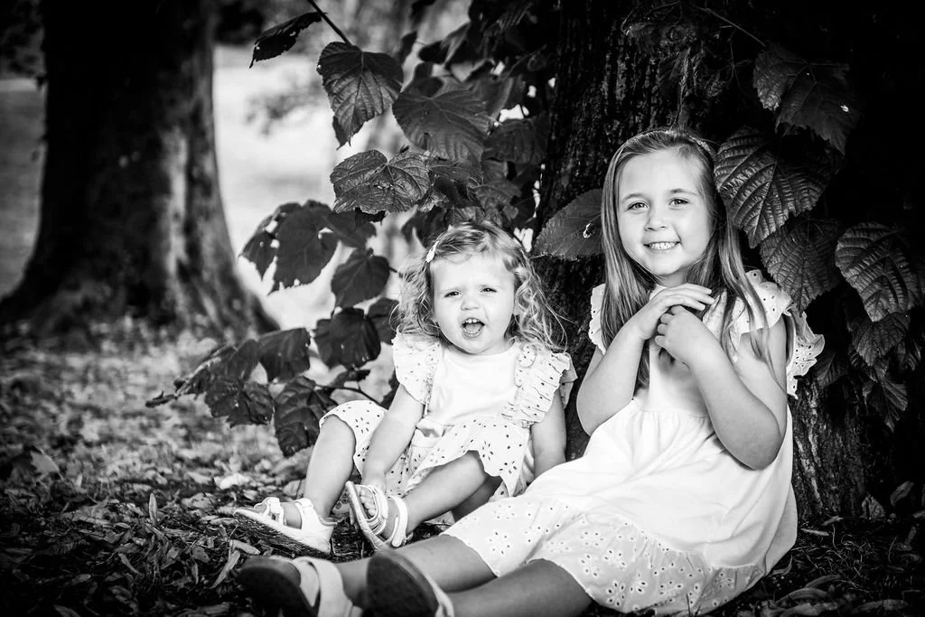 Black and white photograph of two young sisters sitting together in the leaves under a tree during a children's photoshoot at Belle Vue Park, Newport, by Emotion Picture Photography.