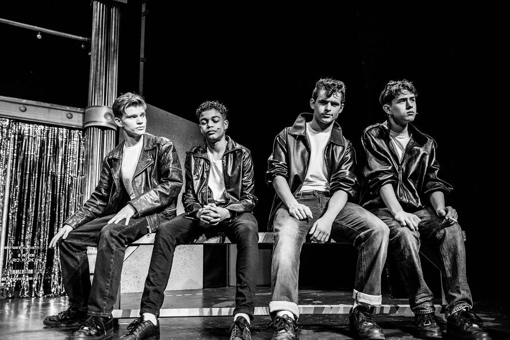 Four boys sitting on a bench on stage, wearing leather jackets and jeans, with a curtain and stage equipment in the background.