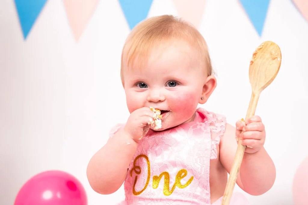 A young girl celebrating her first birthday, wearing a pink dress with the number one on it, holding a wooden spoon, and eating cake with a happy expression.