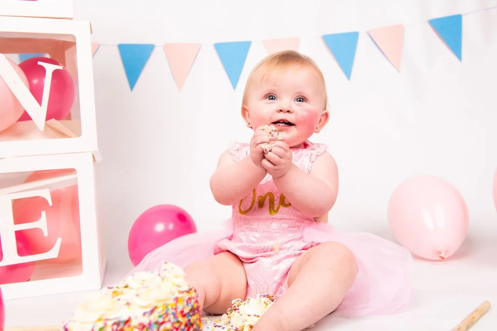 Adorable baby girl celebrating her first birthday, sitting on the floor surrounded by pink balloons, cake, and birthday decorations, wearing a pink dress with 'One' written on it.