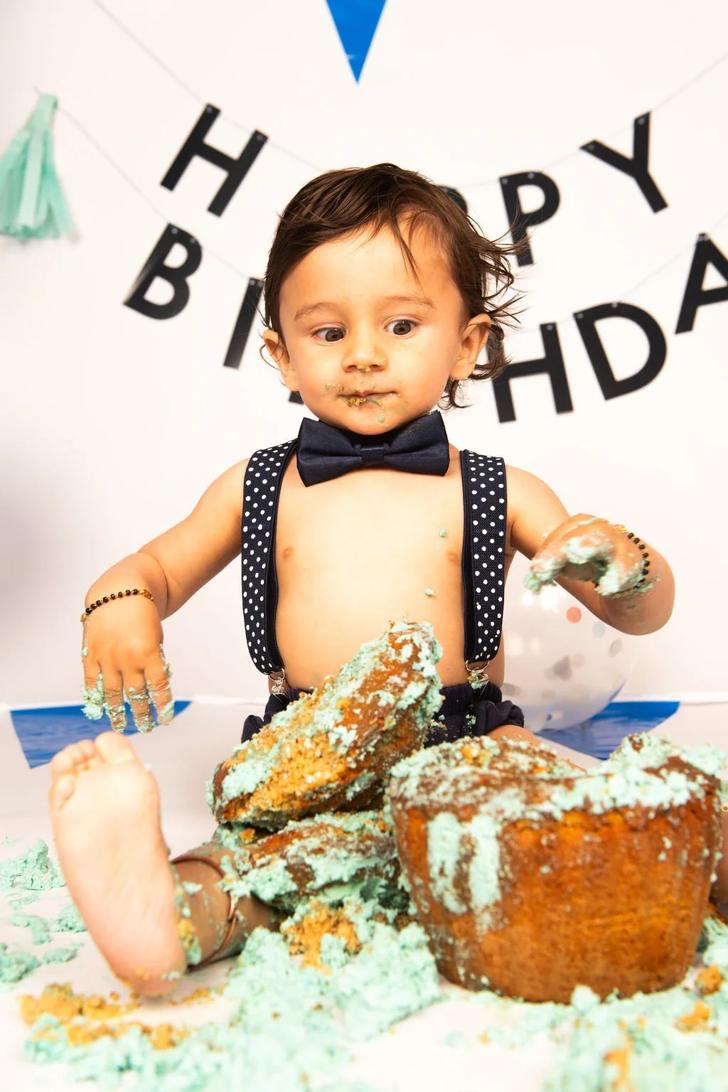 A young boy at a birthday party is covered in green frosting while playing with large pieces of cake on a table. He is shirtless, wearing a bow tie and suspenders, and has messy hair. In the background, a "Happy Birthday" banner is visible.