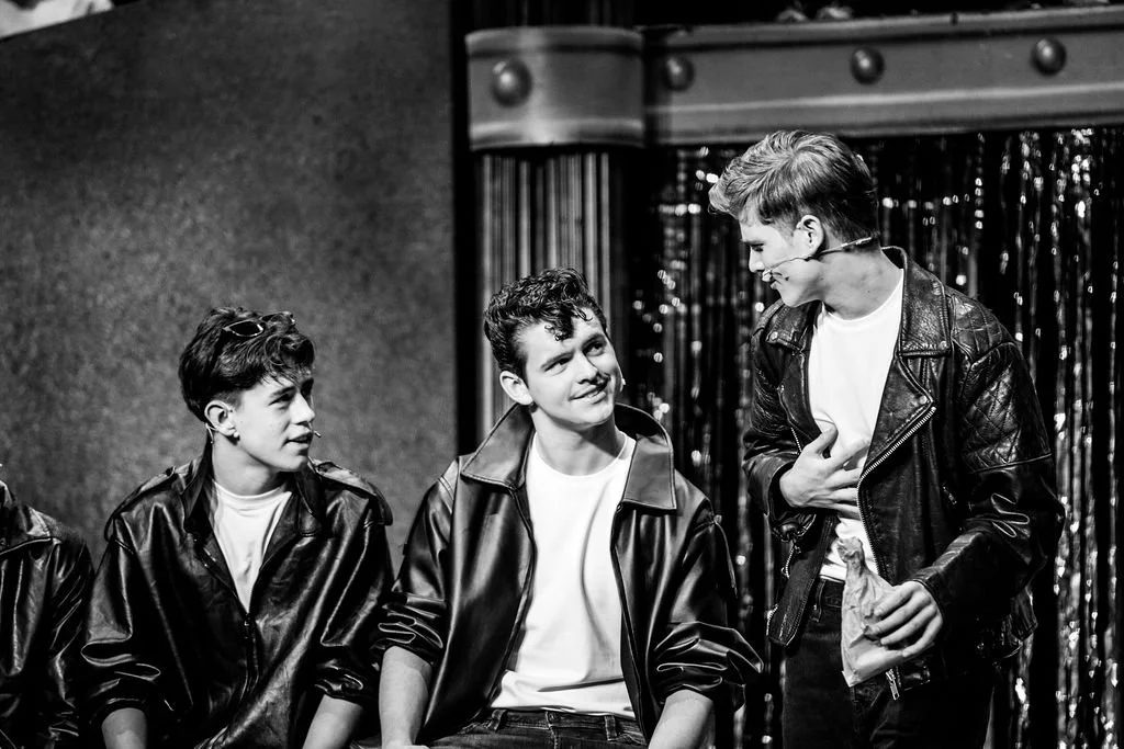 Four young men in leather jackets stand on stage with a curtain behind them, engaging in a conversation. One man is holding a paper bag, and they are smiling and looking at each other.