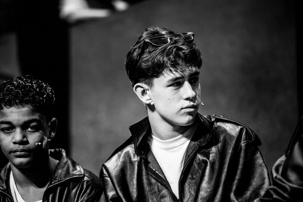 Two young males with short dark hair, one with goggles on head, sit side by side wearing leather jackets, in black and white photo.